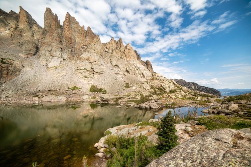 A scenic view of rugged mountains reflecting in a calm lake under a partly cloudy sky. Lush greenery in the foreground.