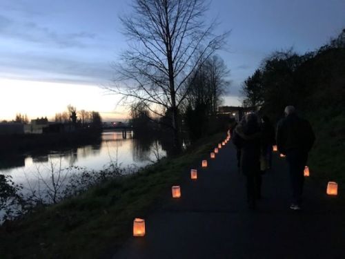 A serene riverside path at dusk, lined with glowing lanterns and people walking along the water's edge.