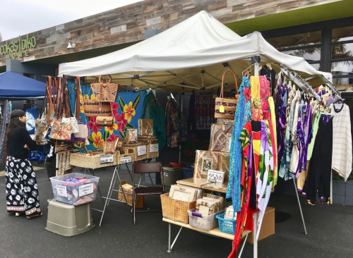 A vibrant market stall with colorful fabrics, bags, and crafts under a canopy, set in an outdoor market.