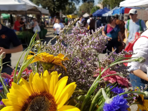 A vibrant bouquet of sunflowers and purple flowers in focus, with a bustling market scene in the background.
