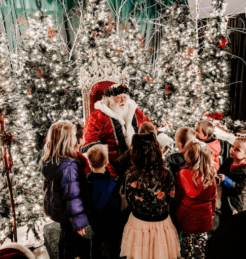 Santa Claus sits on a throne surrounded by children, with decorated Christmas trees and lights in the background.