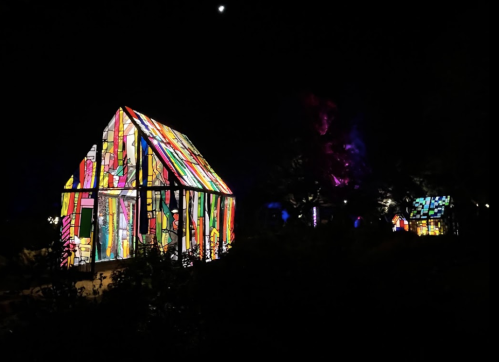 A brightly lit, colorful stained glass house at night, surrounded by trees and a dark sky.