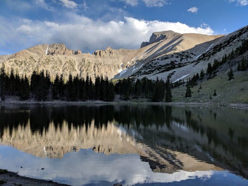 A serene mountain lake reflecting surrounding peaks and trees under a partly cloudy sky.