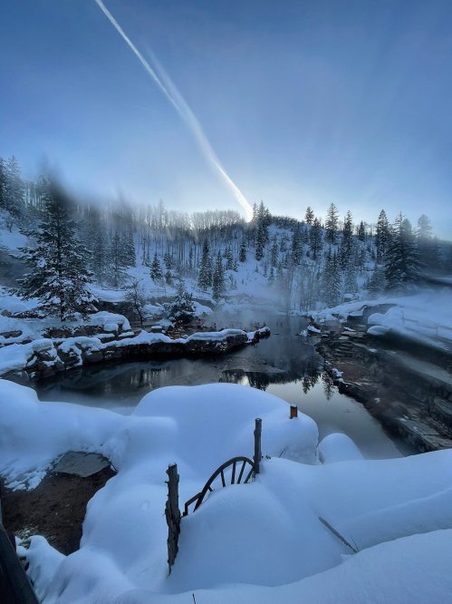 A serene winter landscape with snow-covered trees, a frozen river, and a clear blue sky with a jet trail.