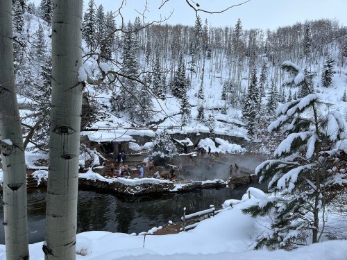A snowy landscape with people enjoying a hot spring, surrounded by trees and mist in a serene winter setting.
