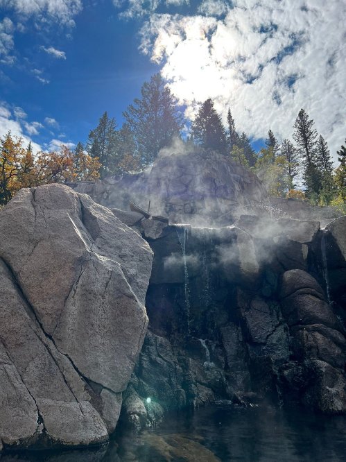 Steam rises from a rocky outcrop near a body of water, surrounded by trees under a bright, cloudy sky.