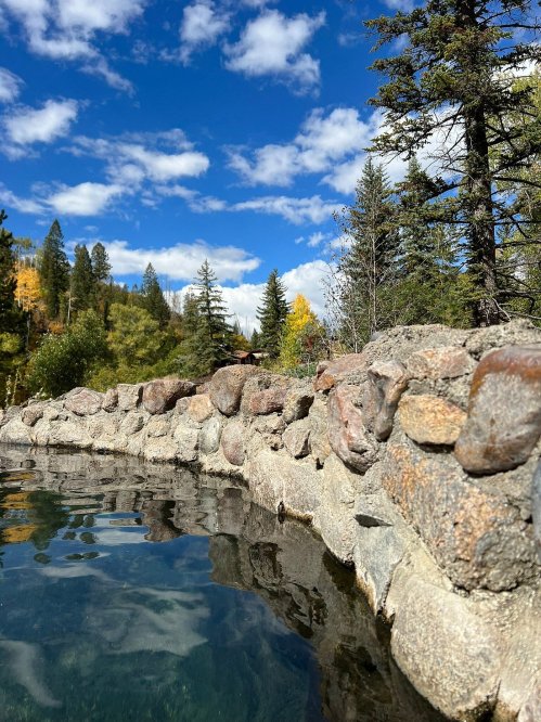 A stone wall beside a clear pool reflects trees and a blue sky with fluffy clouds in a serene outdoor setting.