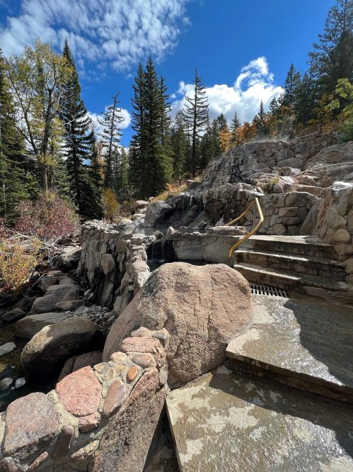 A rocky pathway leads to a natural hot spring, surrounded by trees and a bright blue sky with scattered clouds.