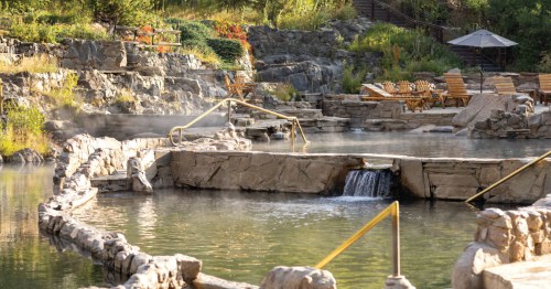 A serene hot spring with stone pathways, gentle waterfalls, and wooden loungers surrounded by lush greenery.