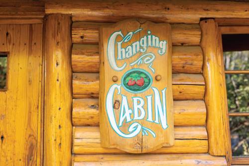 A wooden sign reading "Changing Cabin" hangs on a log cabin wall. The sign features decorative lettering and a small graphic.