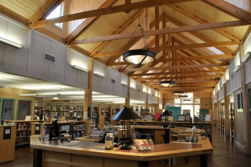 A spacious library interior with wooden beams, bookshelves, and a central checkout desk. Natural light fills the room.