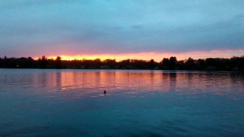 A serene lake at sunset, reflecting vibrant orange and purple hues in the water, with trees lining the shore.