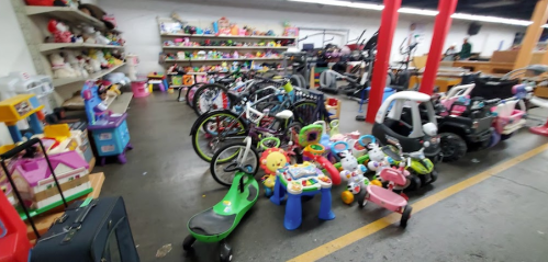 A variety of children's toys and bikes are scattered across a store floor, with shelves of toys in the background.