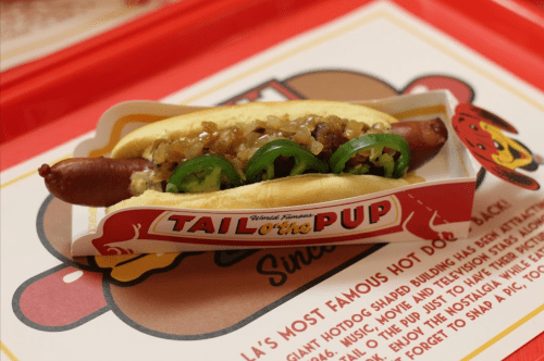 A hot dog in a bun topped with onions and jalapeños, served in a branded paper tray.