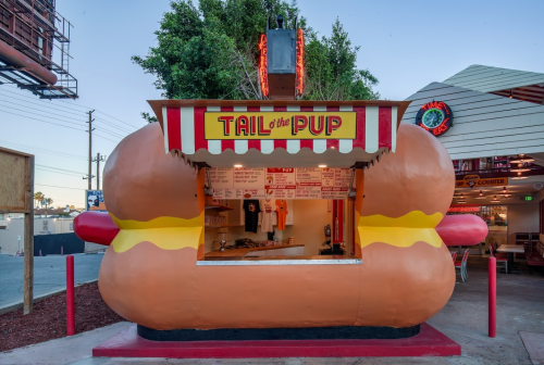 A whimsical hot dog stand shaped like a giant hot dog, featuring a red and white striped awning and neon sign.