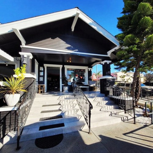 A charming entrance to a dark wooden building with stairs, railings, and palm trees under a clear blue sky.