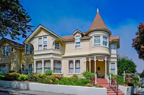 A charming yellow Victorian house with a turret, surrounded by greenery and colorful flowers under a clear blue sky.