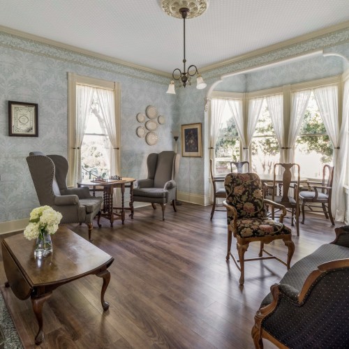 A cozy living room with vintage furniture, large windows, and floral decor, creating a warm and inviting atmosphere.