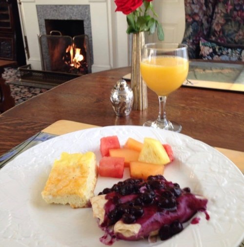 A plate with fruit, a baked dish, and a blueberry dessert, alongside a glass of orange juice and a rose in a vase.