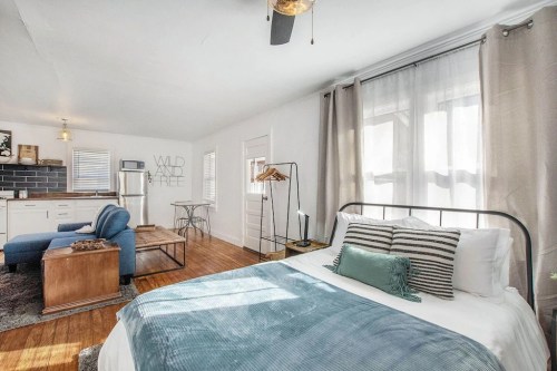 Cozy bedroom with a blue throw, wooden furniture, and a living area featuring a sofa and kitchenette. Bright and inviting space.