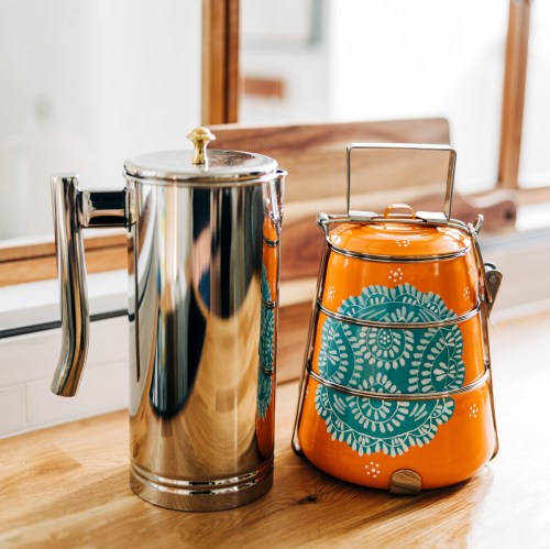 A shiny silver French press next to a vibrant orange tiffin with a floral design, set on a wooden countertop.