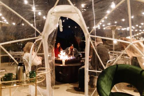 A cozy outdoor gathering in a transparent dome with string lights, featuring people around a fire pit.