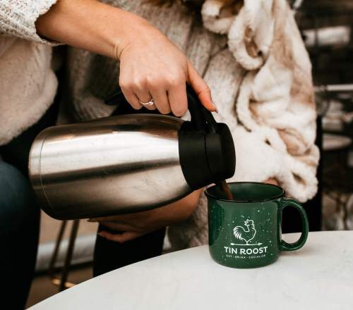 A person pours coffee from a stainless steel thermos into a green mug with "TIN ROOST" printed on it.