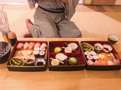 A person in traditional attire sits on a tatami mat, surrounded by three trays of sushi and green tea.
