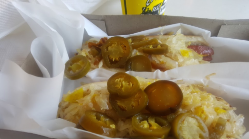 Two hot dogs topped with sauerkraut and sliced jalapeños in a takeout box, with a drink in the background.
