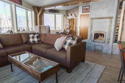 Cozy living room with a brown sectional sofa, a glass coffee table, and a stone fireplace, featuring large windows and mountain views.