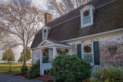 A charming brick house with green shutters and holiday wreaths on the windows, surrounded by trees and greenery.
