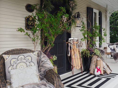 Cozy porch with wicker furniture, greenery, sleds, and a decorative penguin on a striped rug.