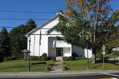 A white town hall building with a sloped roof, surrounded by green grass and trees, under a clear blue sky.