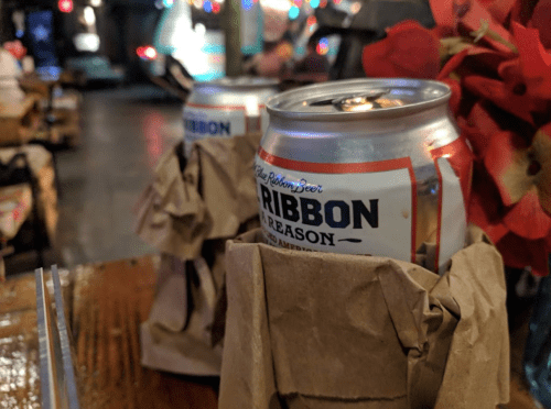 Two cans of beer wrapped in brown paper bags, placed on a wooden table with colorful lights in the background.