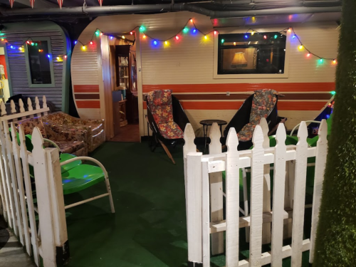 A cozy outdoor space with colorful lights, a white picket fence, and seating around a vintage trailer.