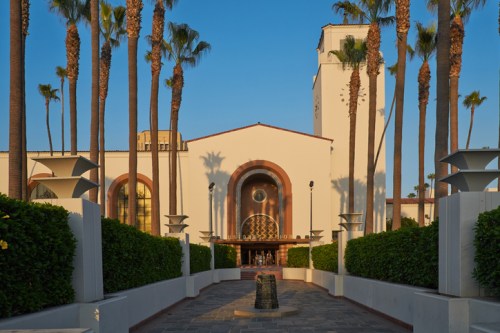 A sunny view of a white building with palm trees, featuring a clock tower and a landscaped pathway leading to the entrance.