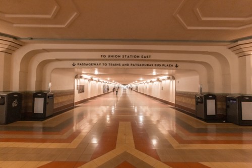 A long, empty corridor in a transit station with signs directing to Union Station East and bus plaza.