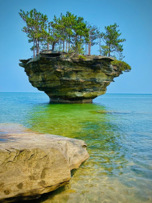 A small island with trees sits atop a rocky formation in clear blue water, under a bright blue sky.