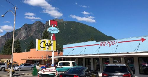 A colorful cafe with a mountain backdrop, featuring a sign that reads "RR2GO!" and vintage cars parked outside.