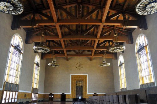 Interior of a grand hall with wooden beams, large windows, and decorative chandeliers, featuring a clock on the wall.