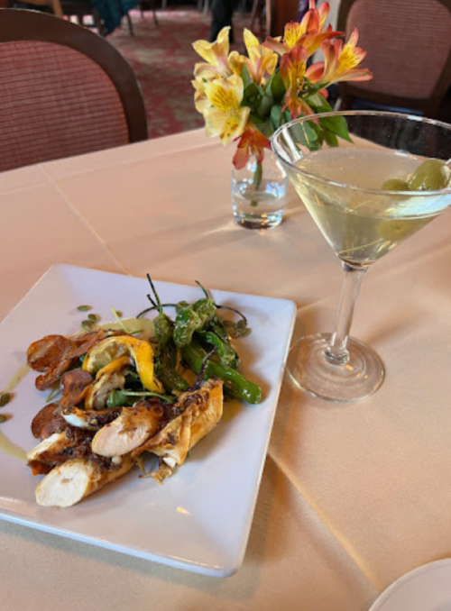 A plate of grilled chicken with vegetables, a martini with olives, and a vase of flowers on a dining table.