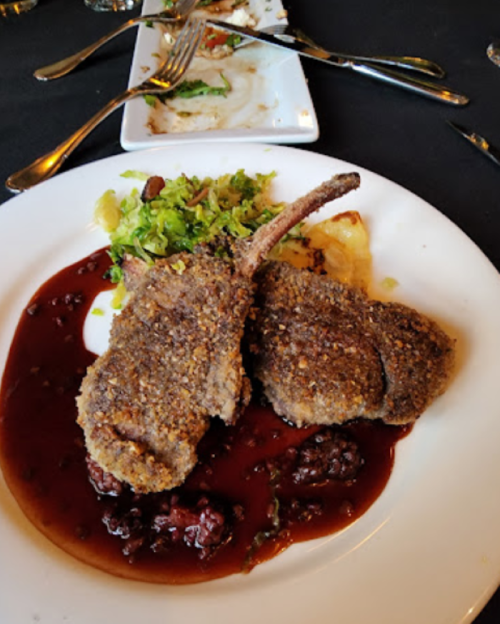 Two breaded lamb chops on a plate with a berry sauce and a side of greens, set on a black tablecloth.