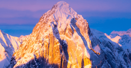Snow-capped mountain peak illuminated by warm sunset light against a clear blue sky.