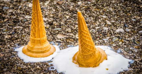 Two ice cream cones tipped over, partially submerged in spilled ice cream on a textured surface.