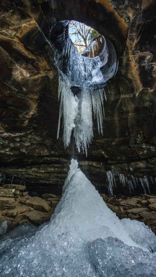 A cave with icicles hanging from the ceiling and a mound of ice on the ground, illuminated by light from above.