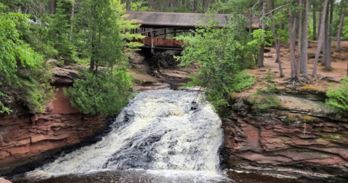 A waterfall cascades over rocky terrain, with a wooden bridge visible in the background, surrounded by lush green trees.