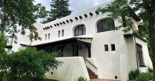 A white, adobe-style house with arched windows and a sloped roof, surrounded by greenery and trees.