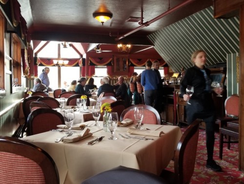 A busy restaurant interior with tables set for dining, patrons chatting, and a server carrying glasses.