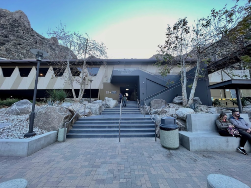 A modern building entrance with stairs, surrounded by rocks and trees, and two people sitting nearby.