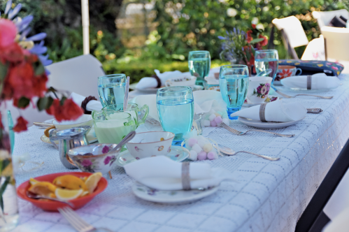 A beautifully set outdoor table with blue glasses, colorful dishes, and floral arrangements, ready for a gathering.